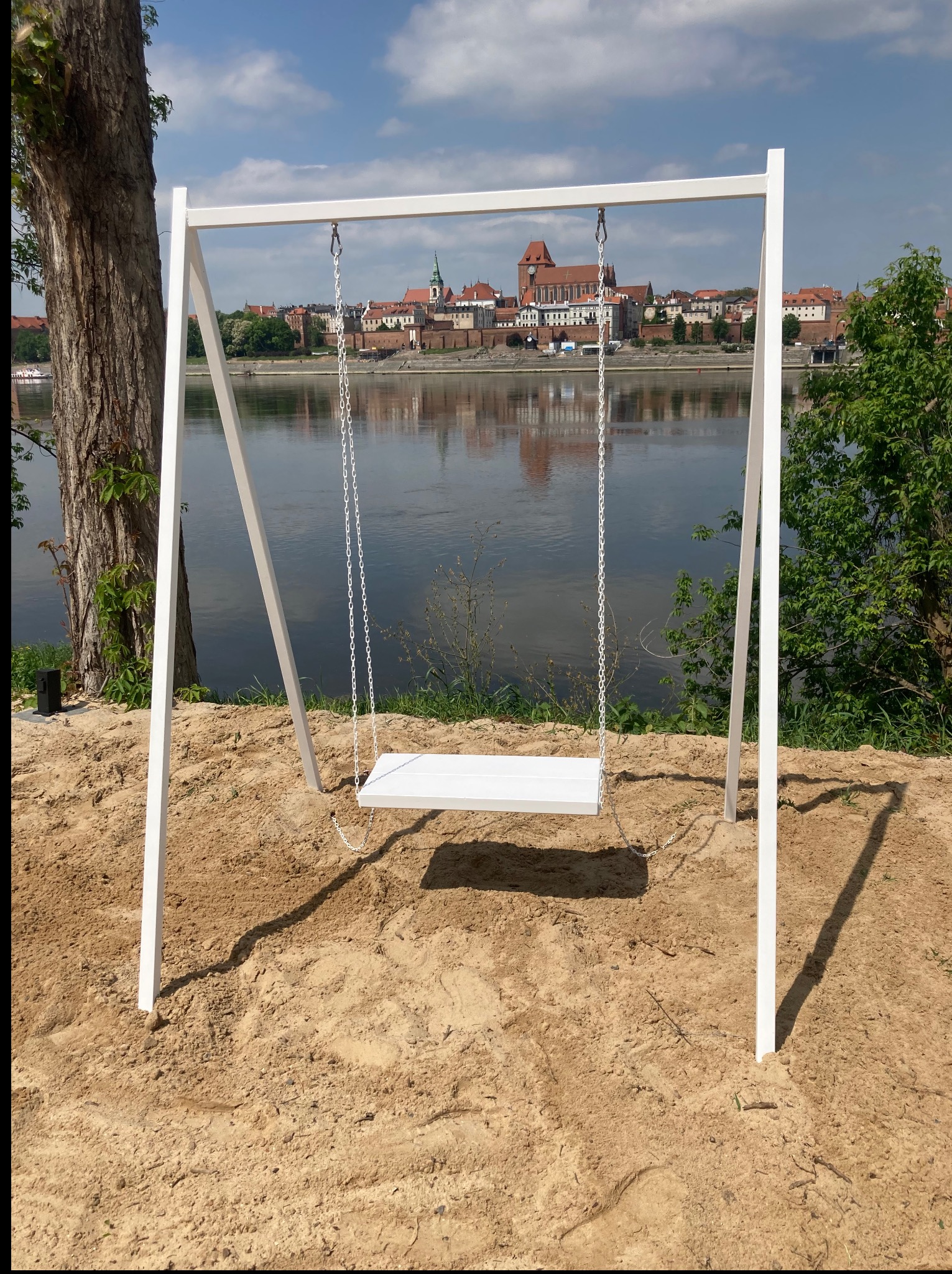 Biała, metalowa huśtawka na łańcuchach, z widokiem na rzekę i zabudowę miejską w tle. Konstrukcja osadzona w piasku, obok drzewo i krzewy.