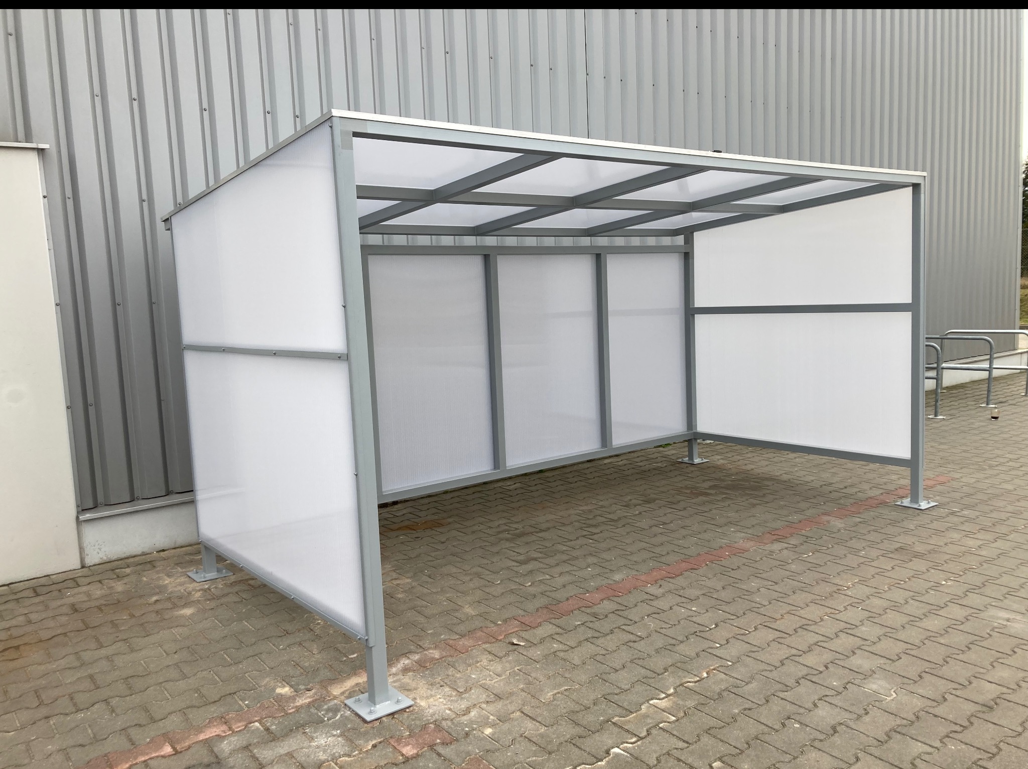 Szara, metalowa konstrukcja z wypełnieniem z tworzywa sztucznego, tworząca zadaszone miejsce postojowe na zewnątrz budynku o szarej elewacji i brukowanym podłożu.