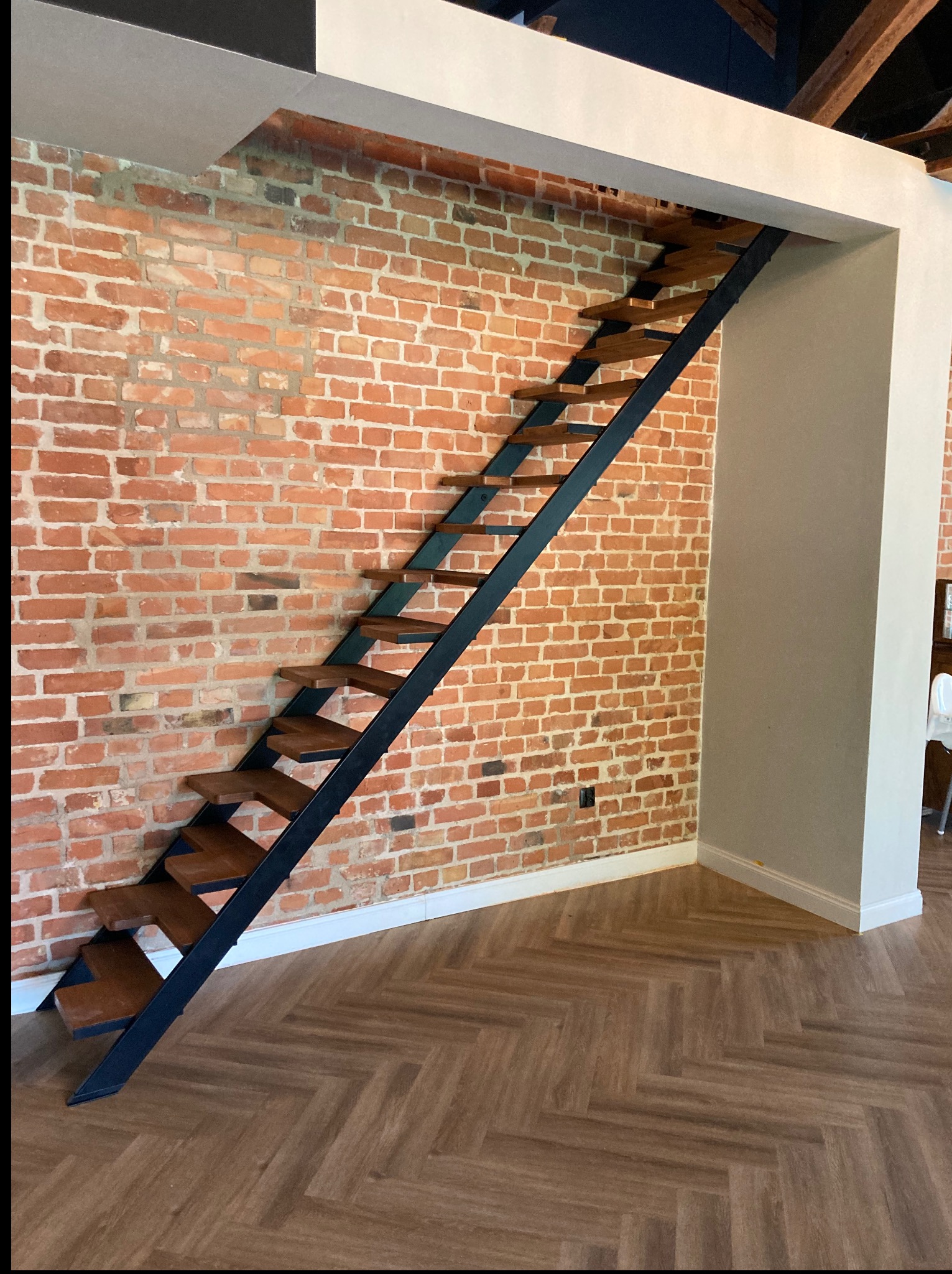 Metalowe schody z drewnianymi stopniami na tle ściany z czerwonej cegły i podłogi w jodełkę. Nowoczesny design, minimalistyczny styl, dobrze oświetlone wnętrze.
