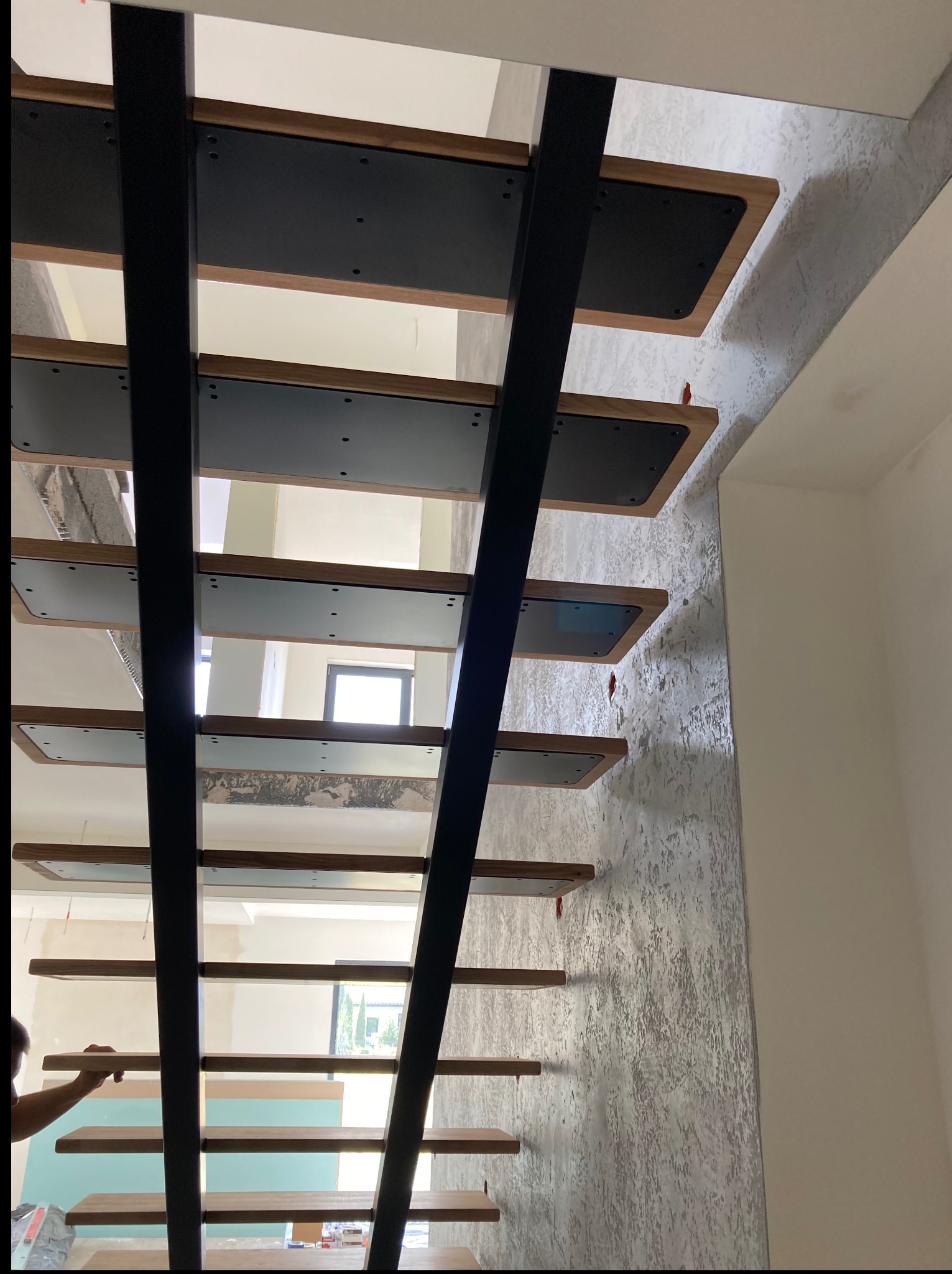 Nowoczesne schody z czarną metalową konstrukcją i drewnianymi stopniami, widok z dołu, podczas montażu, z widocznym fragmentem ściany o dekoracyjnej fakturze.