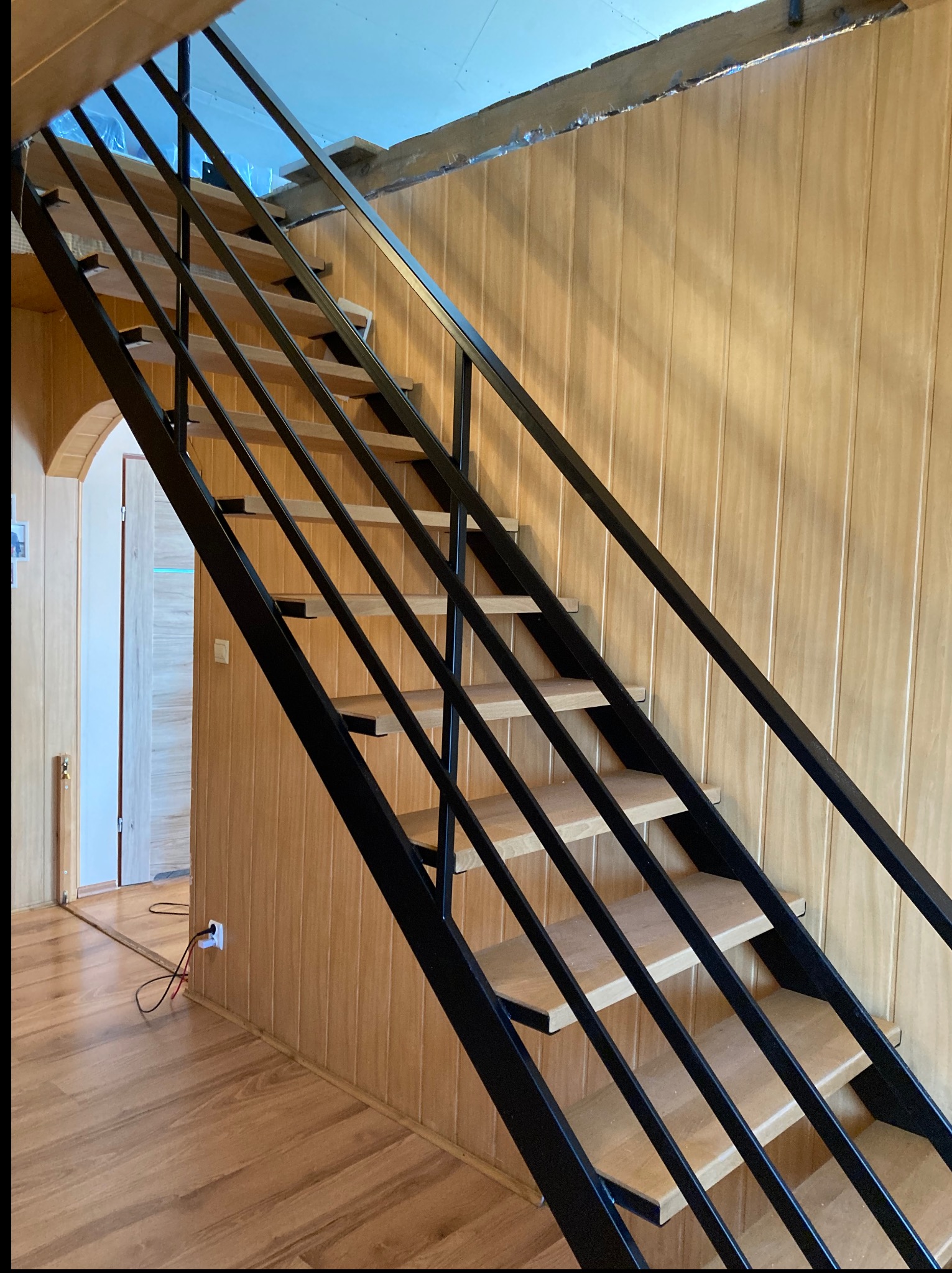 Nowoczesne schody z czarną, metalową konstrukcją i drewnianymi stopniami, widziane z dołu. Jasne drewno na ścianach i podłodze. Widok na wnętrze przez łukowe przejście.