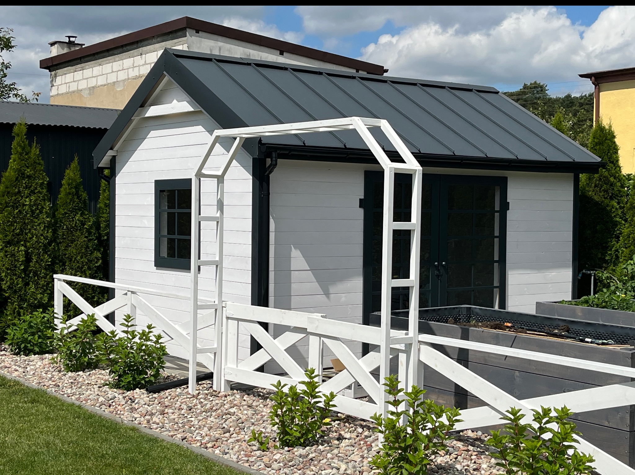 Biała altana z szarą, metalową konstrukcją dachu, otoczona ozdobnym, niskim płotkiem, kamieniami i zielenią. Ujęcie w słoneczny dzień.
