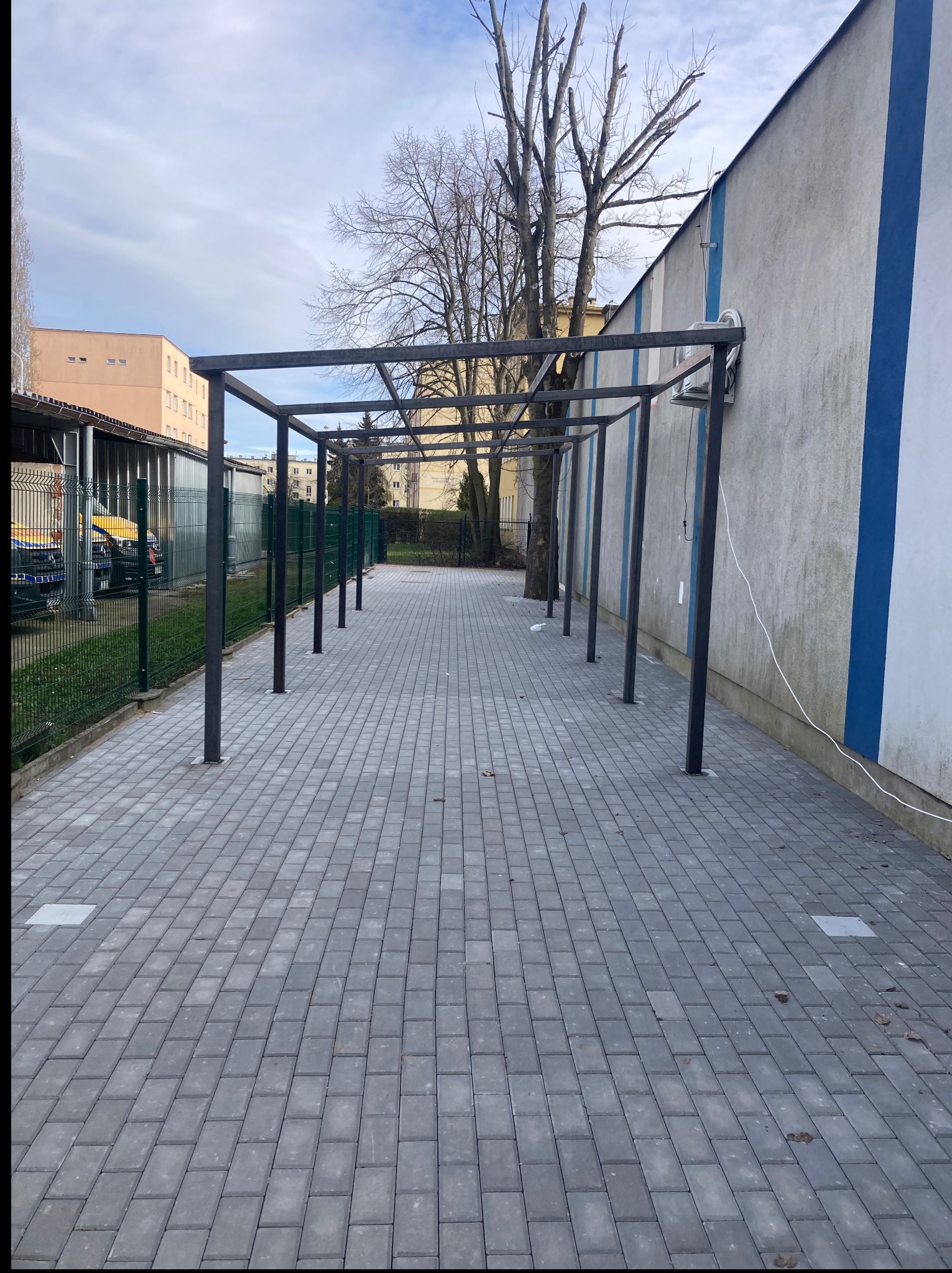 Metalowa konstrukcja pergoli wzdłuż budynku z niebieskim pasem. Szara kostka brukowa na chodniku. Widoczne drzewa i budynki w tle. Słoneczny dzień.