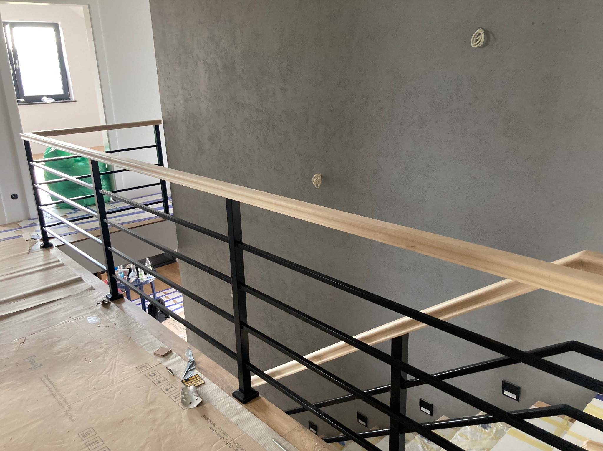 Nowoczesne schody z czarną metalową balustradą i drewnianym poręczem, widok z góry. Podłoga zabezpieczona kartonem.