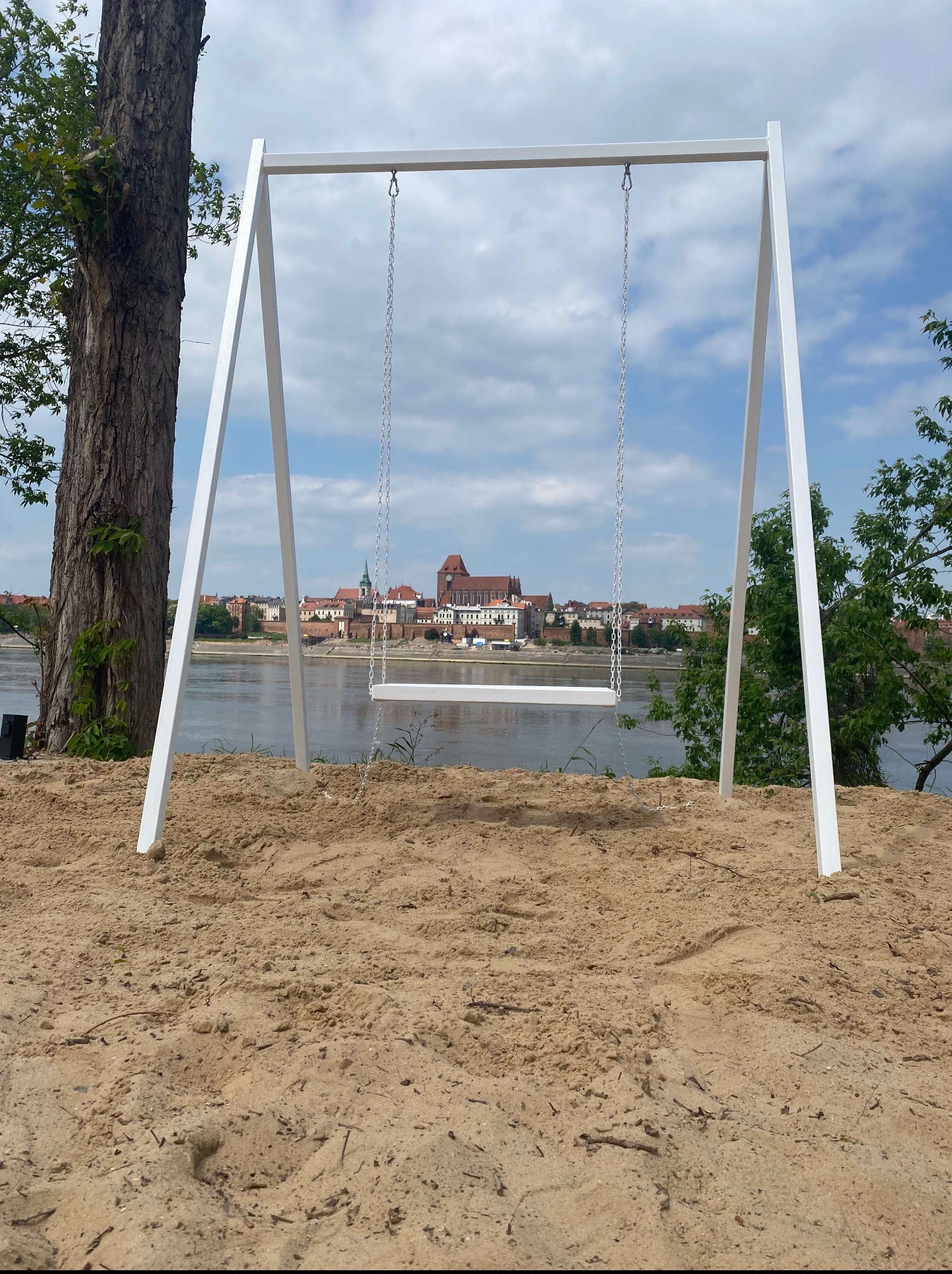 Biała, minimalistyczna huśtawka z metalową ramą na piaszczystym podłożu, z widokiem na rzekę i zabudowę miejską w tle, w słoneczny dzień.