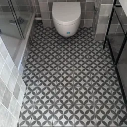 Wykończona łazienka z szaro-białymi płytkami podłogowymi w geometryczny wzór, widoczny fragment kabiny prysznicowej i toaleta. Minimalistyczny design.