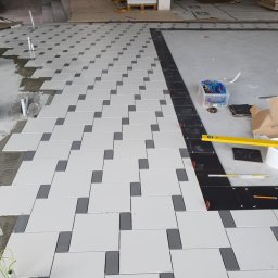 Glazkam - Układanie wzoru z białych i szarych płytek na podłodze, etap prac. Widoczny poziomica, pudełko z narzędziami glazurniczymi i czarne płytki na obrzeżach.