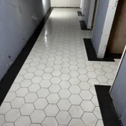 Nowo położona podłoga z heksagonalnych, białych płytek z czarną fugą i czarnym pasem przy ścianie w korytarzu. Widoczne kable wystające ze ściany.
