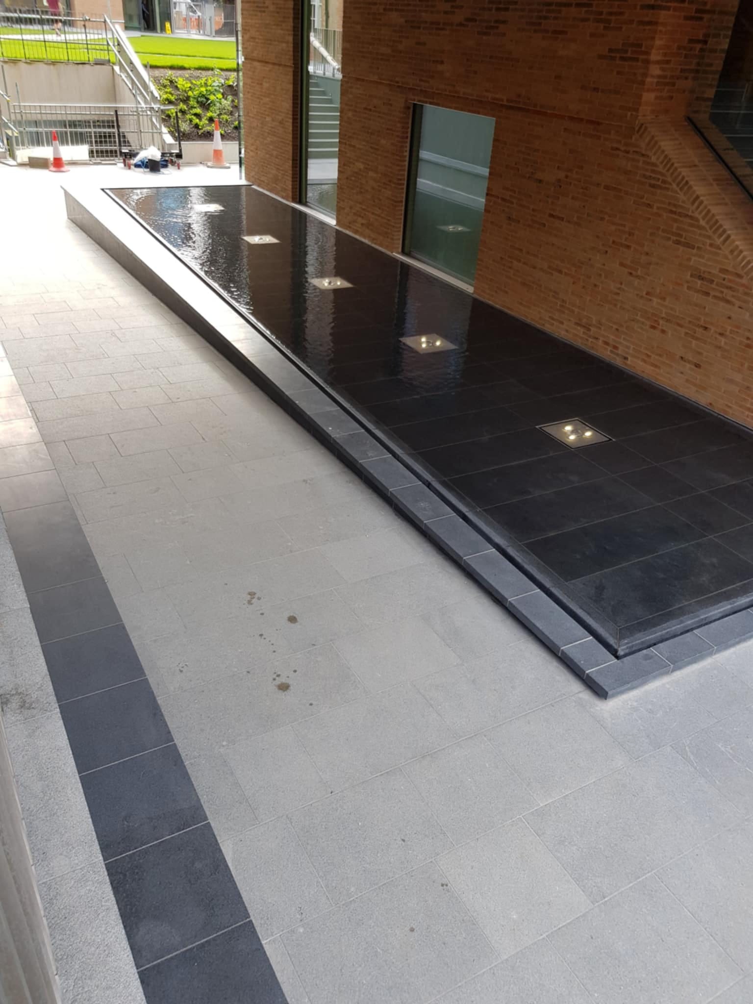 Nowoczesna fontanna z czarnych płytek z wbudowanym oświetleniem, otoczona jasnoszarą kostką brukową i ceglaną ścianą budynku w tle.