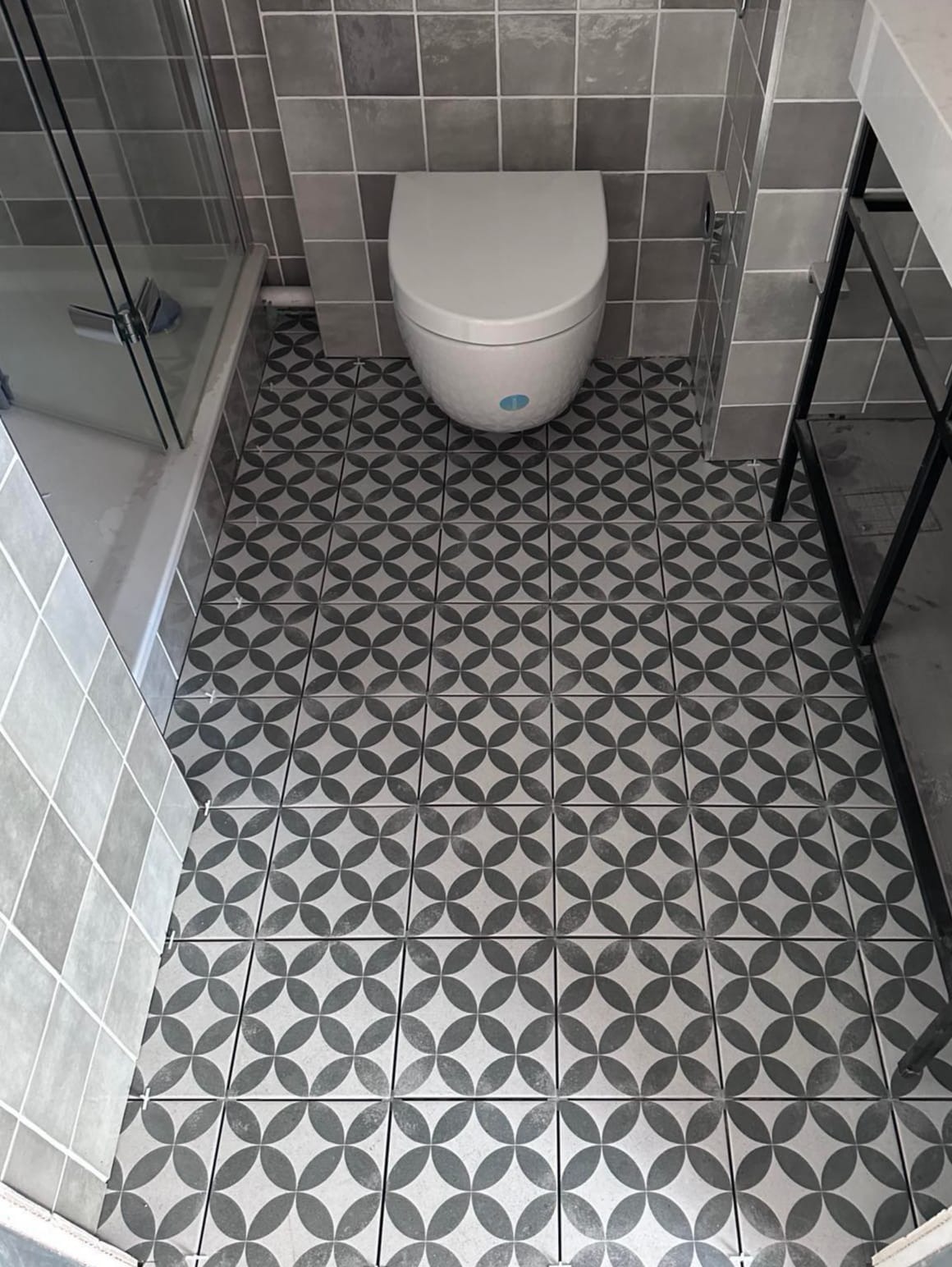 Wykończona łazienka z szaro-białymi płytkami podłogowymi w geometryczny wzór, widoczny fragment kabiny prysznicowej i toaleta. Minimalistyczny design.