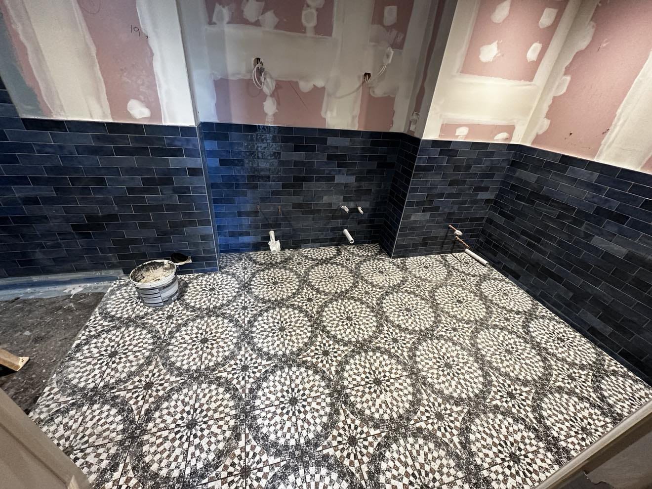 Łazienka w trakcie wykańczania: ułożona podłoga z wzorem, ciemne płytki na ścianach do połowy wysokości, widoczne instalacje i wiadro z zaprawą.