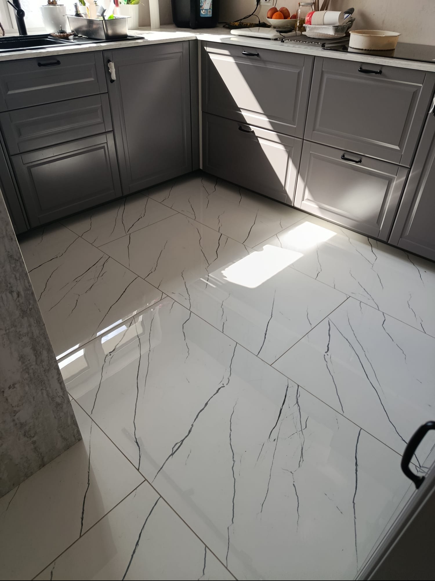 Błyszczące, białe płytki podłogowe z czarnym wzorem żyłek w kuchni z szarymi szafkami. Ujęcie z niskiego kąta, podkreślające wzór podłogi i wpadające światło.
