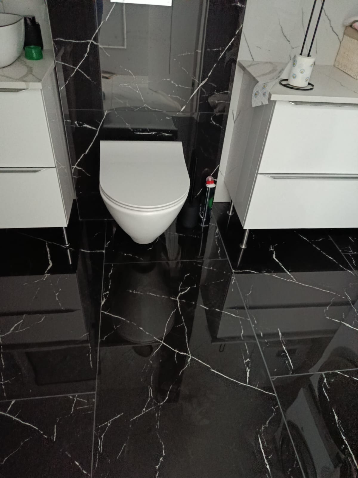 Nowoczesna łazienka z czarnymi płytkami imitującymi marmur, białą wiszącą toaletą i szafkami pod umywalkę. Minimalistyczny design, eleganckie wykończenie.