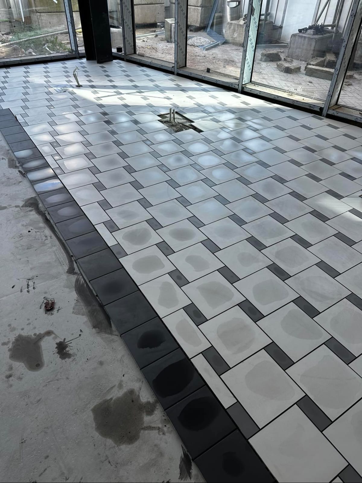 Układanie podłogi z płytek w geometryczny wzór. Widoczne czarne obramowanie i jasne płytki w trakcie montażu, z elementami instalacji wystającymi z podłoża.