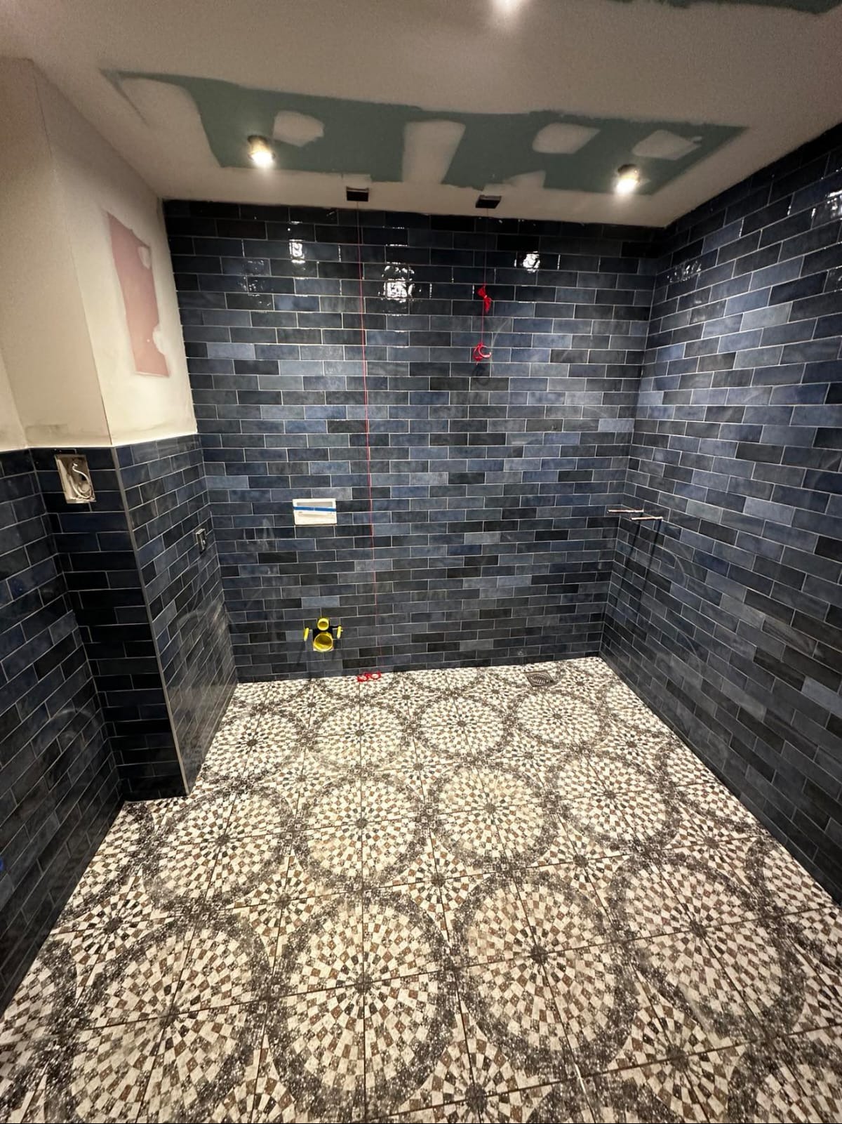 Wykończona łazienka z ciemnymi, błyszczącymi płytkami na ścianach i wzorzystą mozaiką na podłodze. Widoczne elementy instalacji i oświetlenie sufitowe.