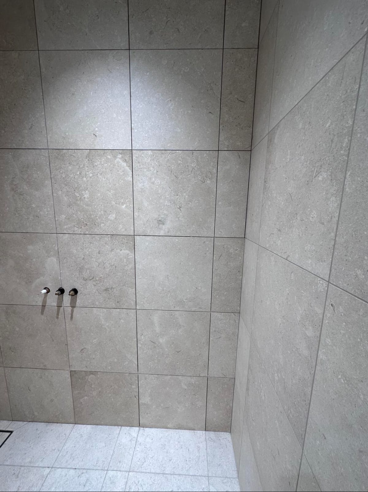 Wykończona ściana i podłoga w łazience, wyłożone jasnymi, kwadratowymi płytkami o marmurkowej strukturze. Widoczne przyłącza wodne.