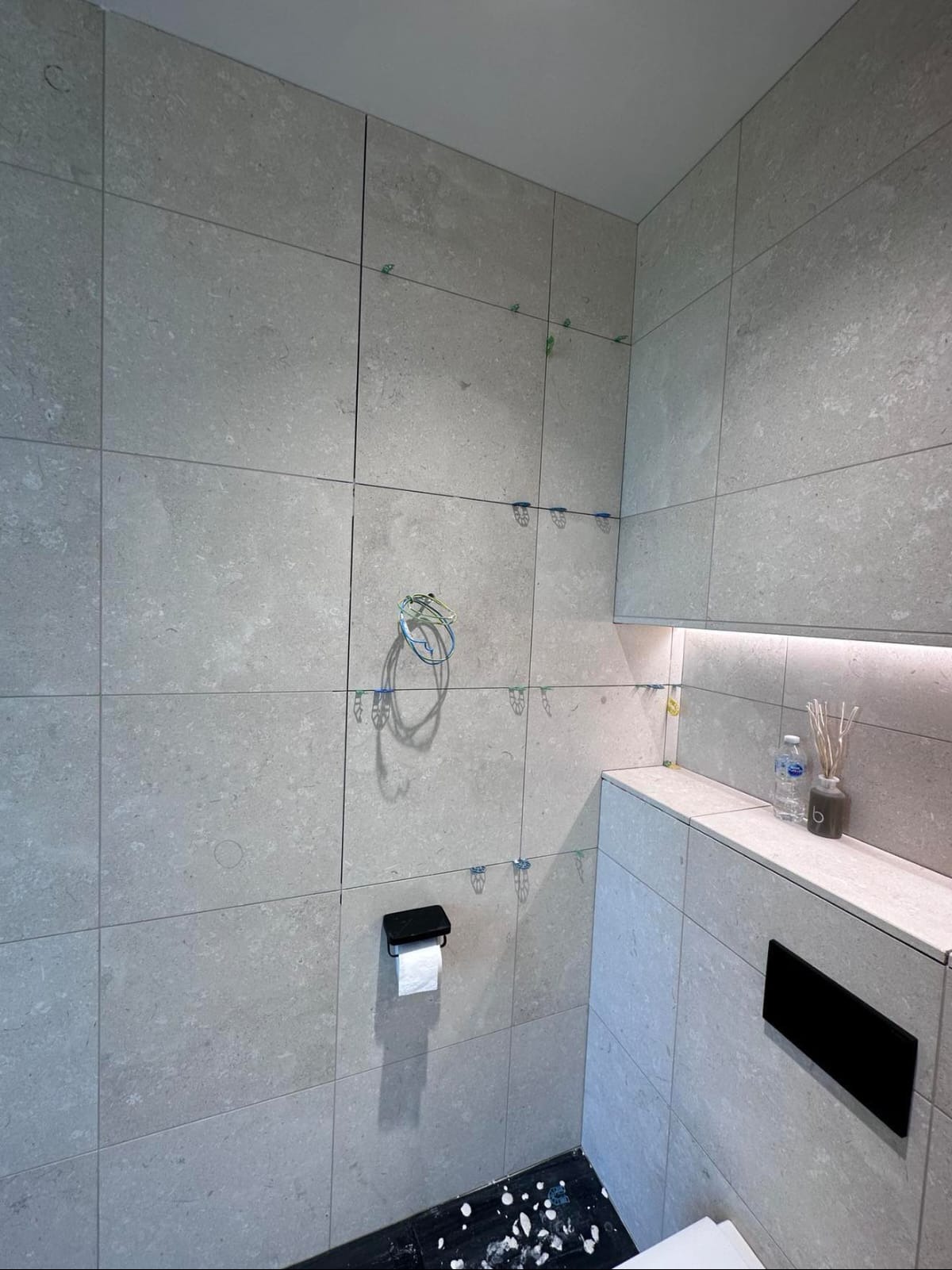 Łazienka w trakcie wykańczania: jasnoszare płytki na ścianach, widoczne elementy instalacji, uchwyt na papier toaletowy i dekoracje na półce.