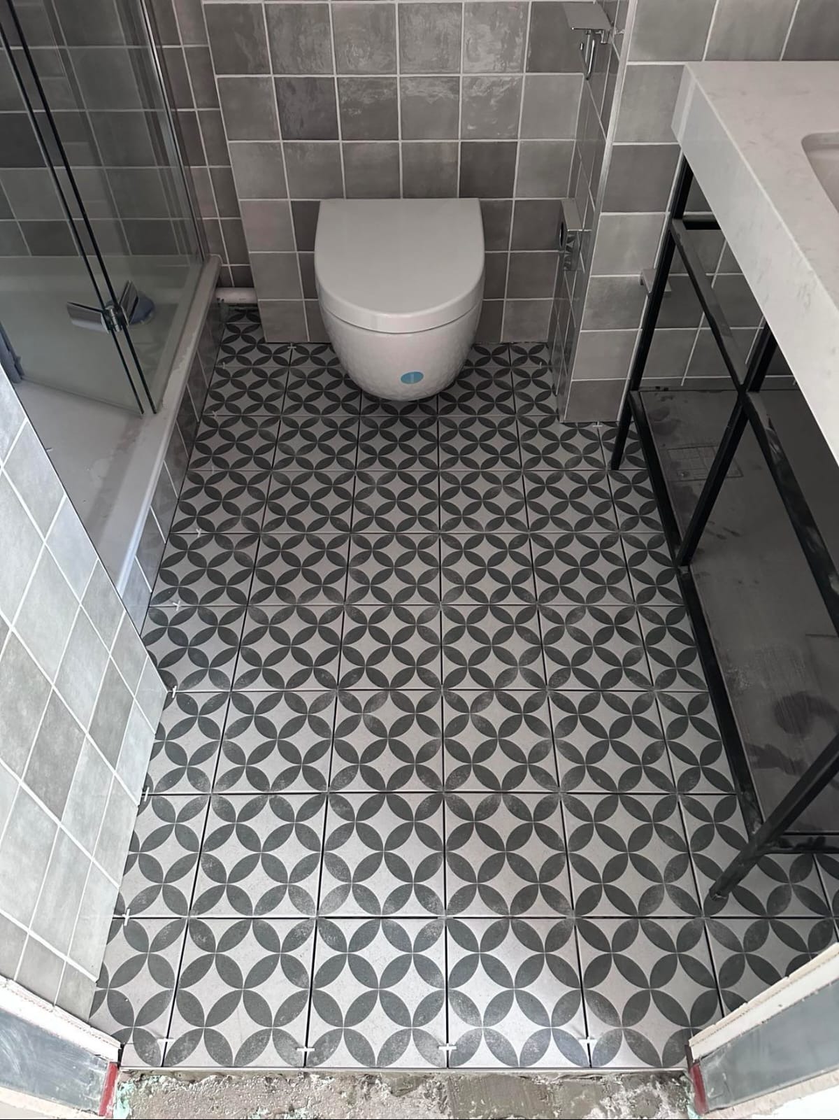 Wykończona łazienka z szaro-białą mozaiką na podłodze i ścianach. Widoczna toaleta, kabina prysznicowa i fragment umywalki. Minimalistyczny, nowoczesny styl.