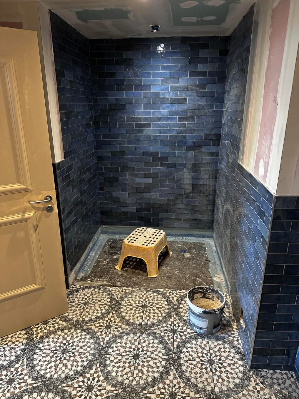 Łazienka w trakcie prac: ściany wyłożone granatową, połyskującą glazurą, widoczny stołek i wiadro z zaprawą na podłodze z wzorzystymi płytkami.