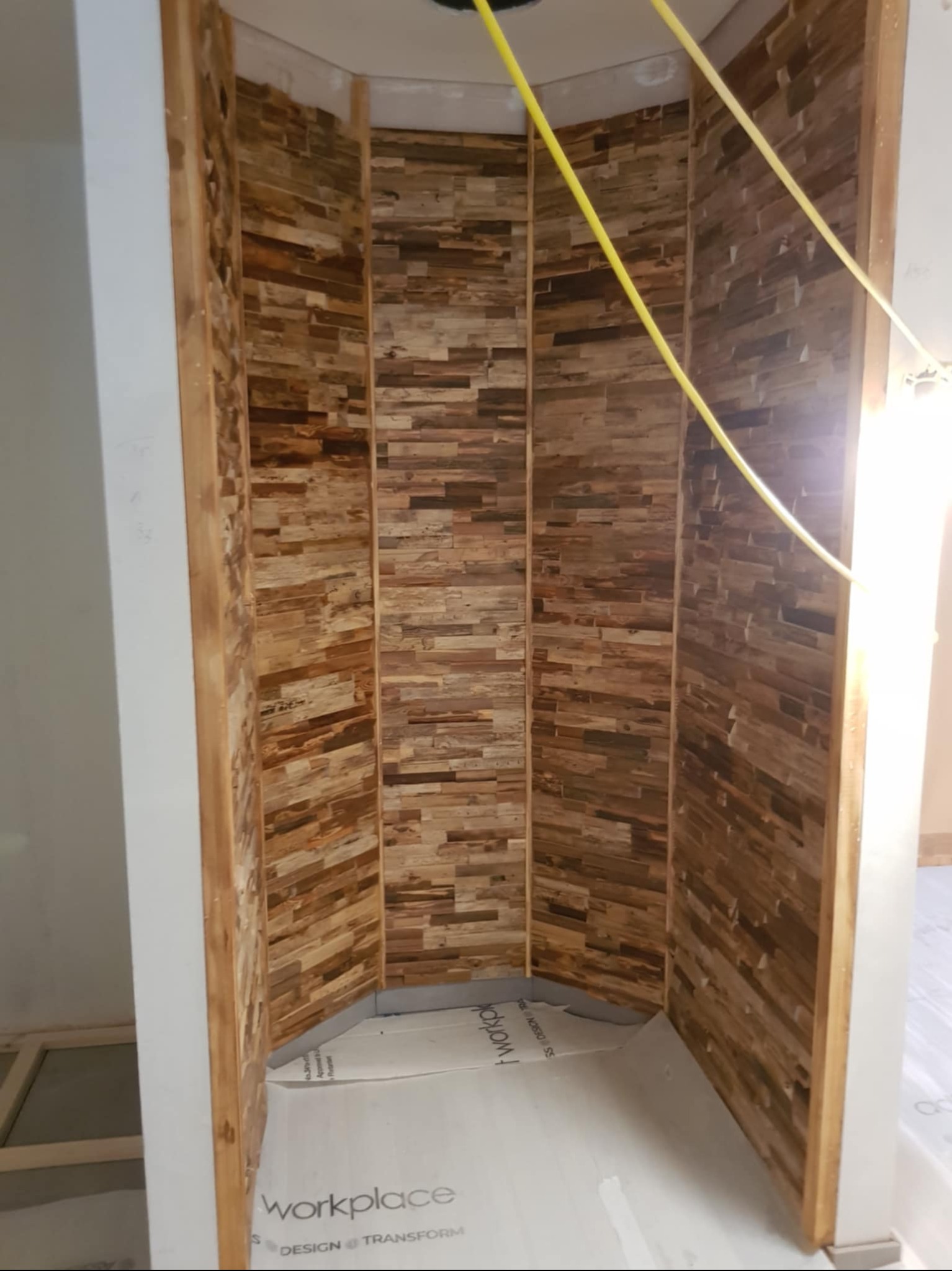Narożna ściana wykończona dekoracyjną okładziną z drewnopodobnych płytek w odcieniach brązu, z widocznym fragmentem instalacji elektrycznej i zabezpieczeniem podłogi.
