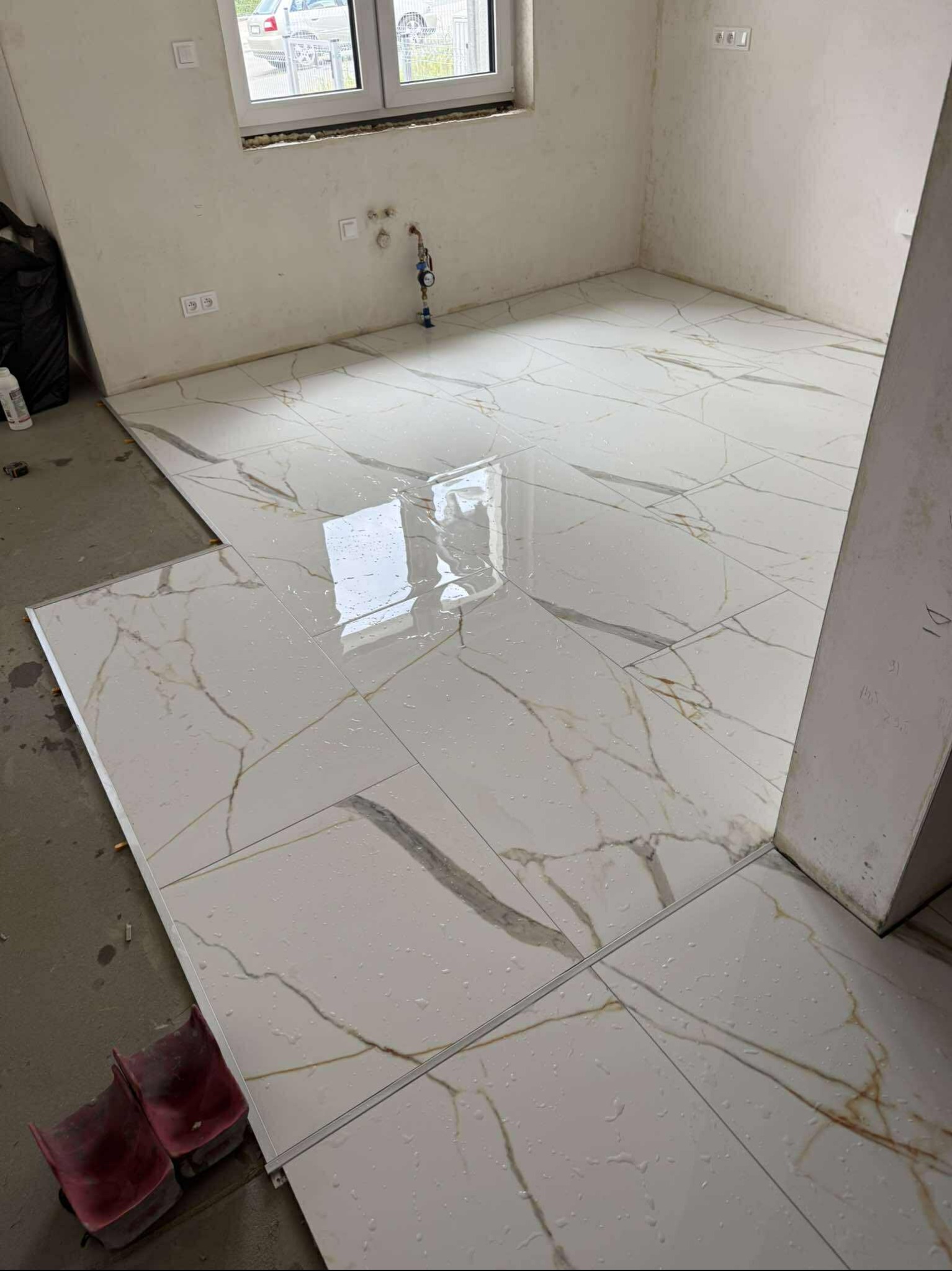 Układanie białych płytek podłogowych z szarym i złotym wzorem w nowym wnętrzu. Widoczne narzędzia glazurnicze i resztki wody na powierzchni płytek.