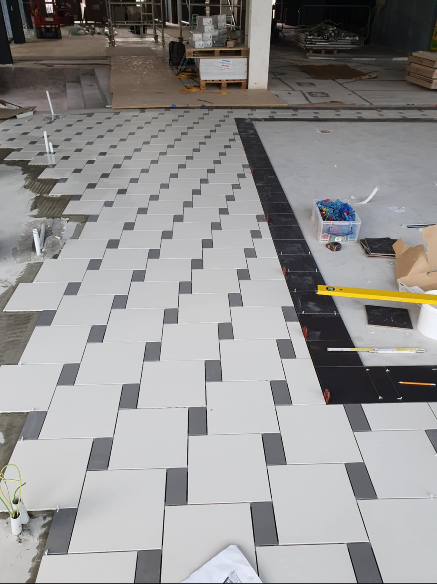 Układanie wzoru z białych i szarych płytek na podłodze, etap prac. Widoczny poziomica, pudełko z narzędziami glazurniczymi i czarne płytki na obrzeżach.