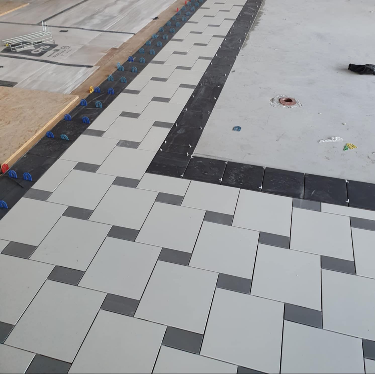 Układanie jasnych płytek podłogowych z ciemnymi akcentami, z użyciem systemów poziomowania. Widoczne elementy instalacji i narzędzia glazurnicze.