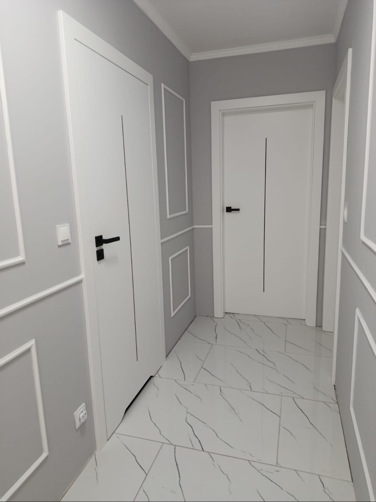 Korytarz w szarościach z białymi drzwiami i marmurową posadzką. Ściany zdobione sztukaterią, minimalistyczne czarne klamki. Nowoczesne wykończenie wnętrza.