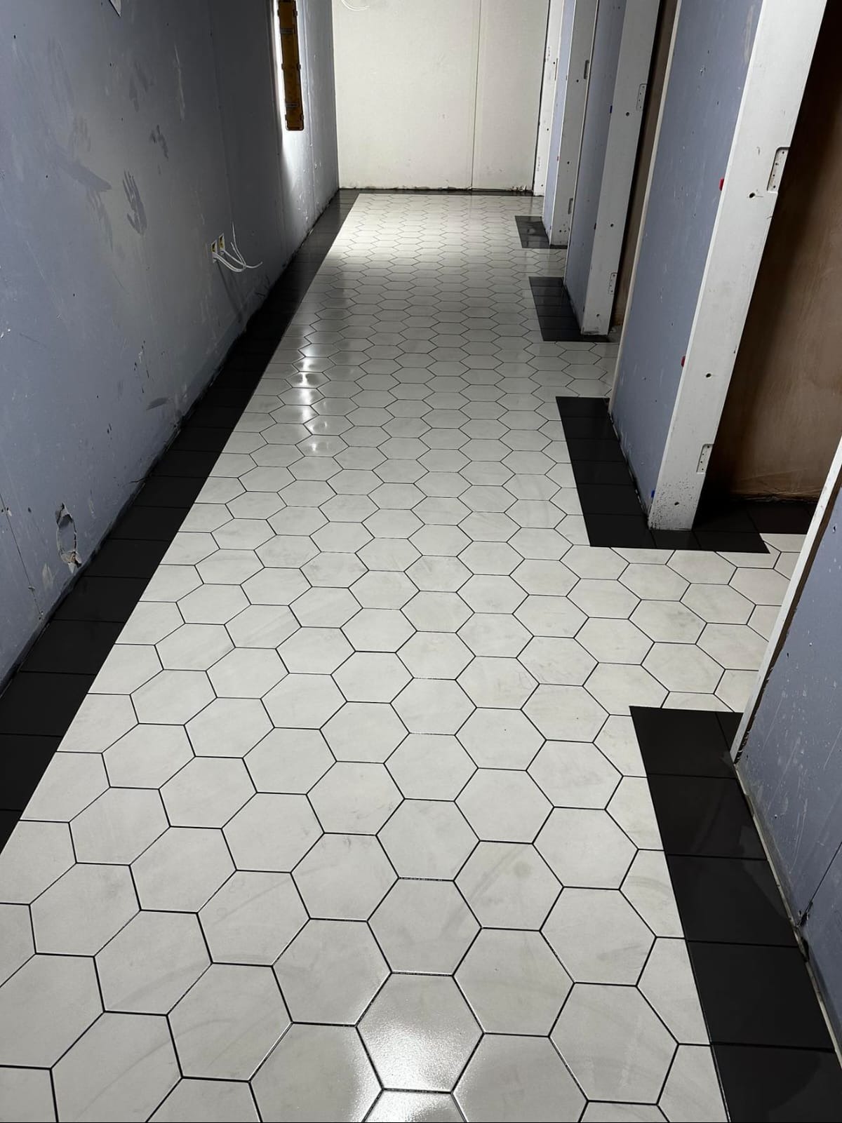 Nowo położona podłoga z heksagonalnych, białych płytek z czarną fugą i czarnym pasem przy ścianie w korytarzu. Widoczne kable wystające ze ściany.