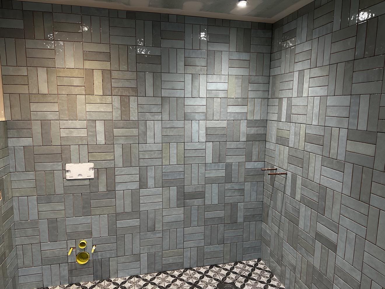 Wykończona łazienka z szaro-niebieskimi płytkami ułożonymi w wzór cegiełki na ścianach i mozaiką na podłodze. Widoczne elementy instalacji wodnej i odpływ liniowy.