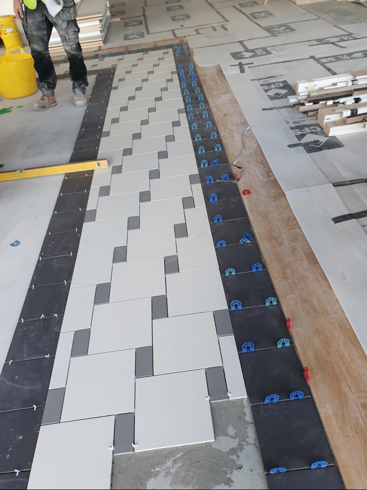 Układanie wzoru z płytek: czarne obramowanie, białe i szare kwadraty w zygzak, poziomica, system poziomowania. Widok z góry na podłogę w trakcie prac.