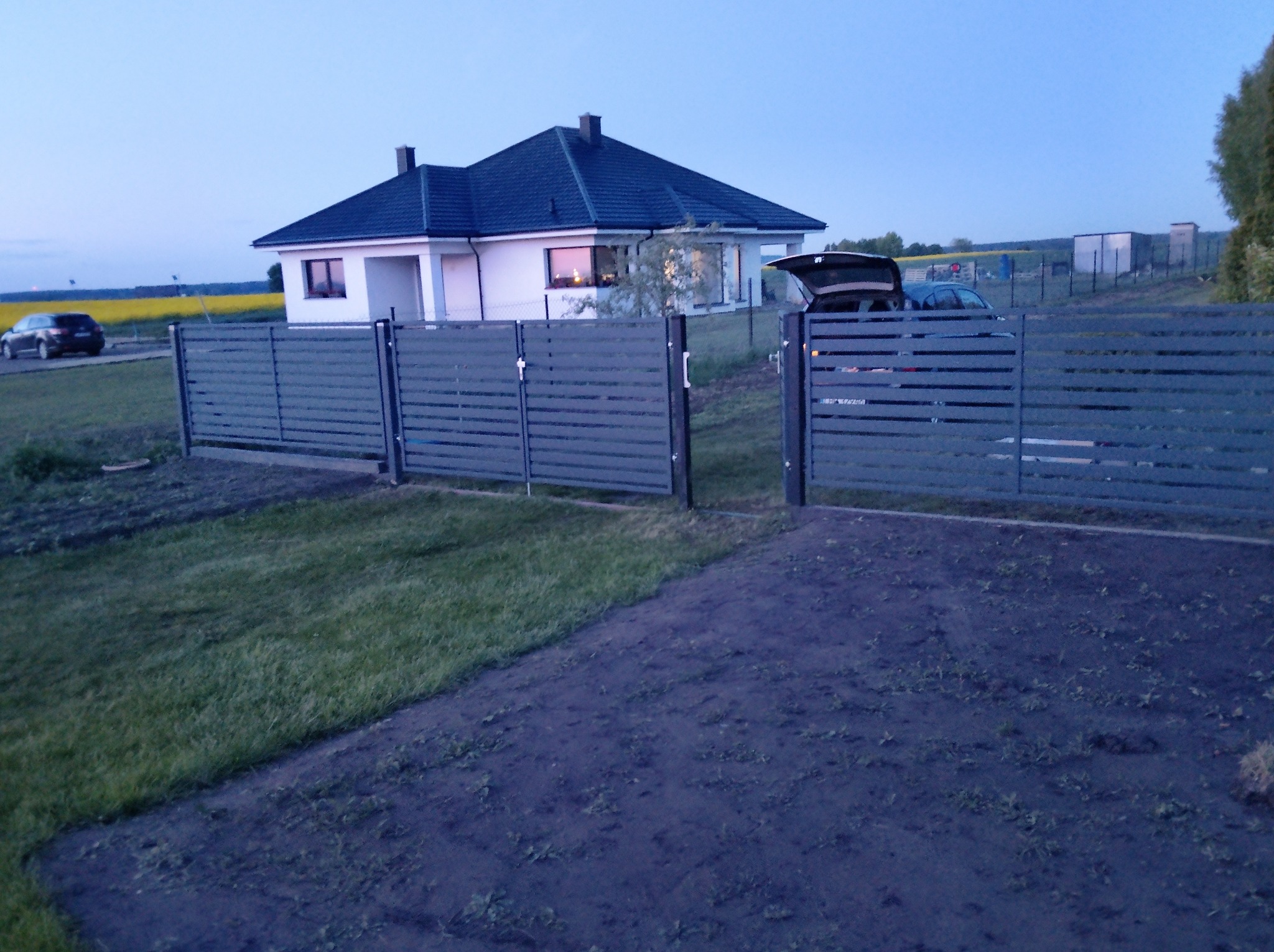 Nowoczesne, poziome ogrodzenie panelowe w kolorze szarym, z widocznym domem jednorodzinnym w tle i samochodem z otwartym bagażnikiem. Ujęcie z dołu, wieczorne światło.