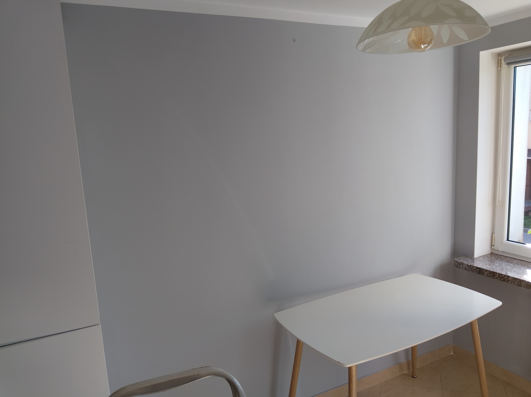 Szara ściana po malowaniu, biały stół z drewnianymi nogami, okno z kamiennym parapetem, lampa sufitowa. Minimalizm, neutralna kolorystyka, gładka powierzchnia ściany.