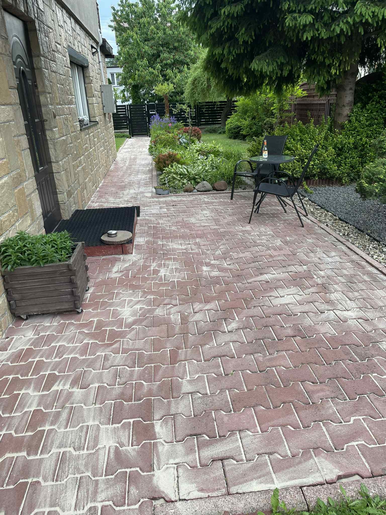 Czerwona kostka brukowa przed domem z kamienną elewacją, stolik ogrodowy z butelką. Widok na uporządkowany ogród z roślinami i czarnym płotem.