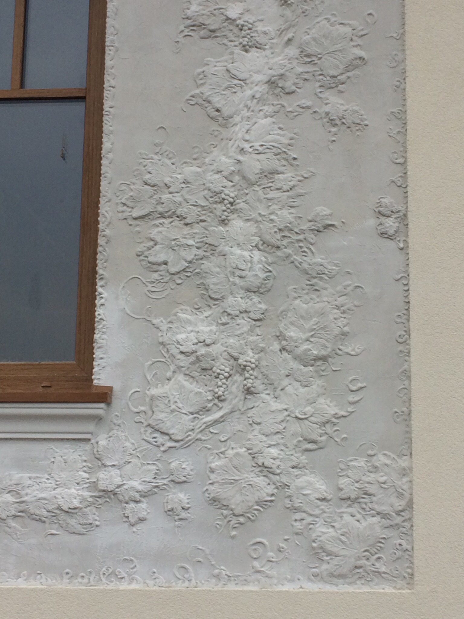 Dekoracyjna elewacja budynku z motywem winorośli, wykonana w jasnych odcieniach, sąsiadująca z oknem o drewnianej ramie. Wyjątkowa faktura i detale.