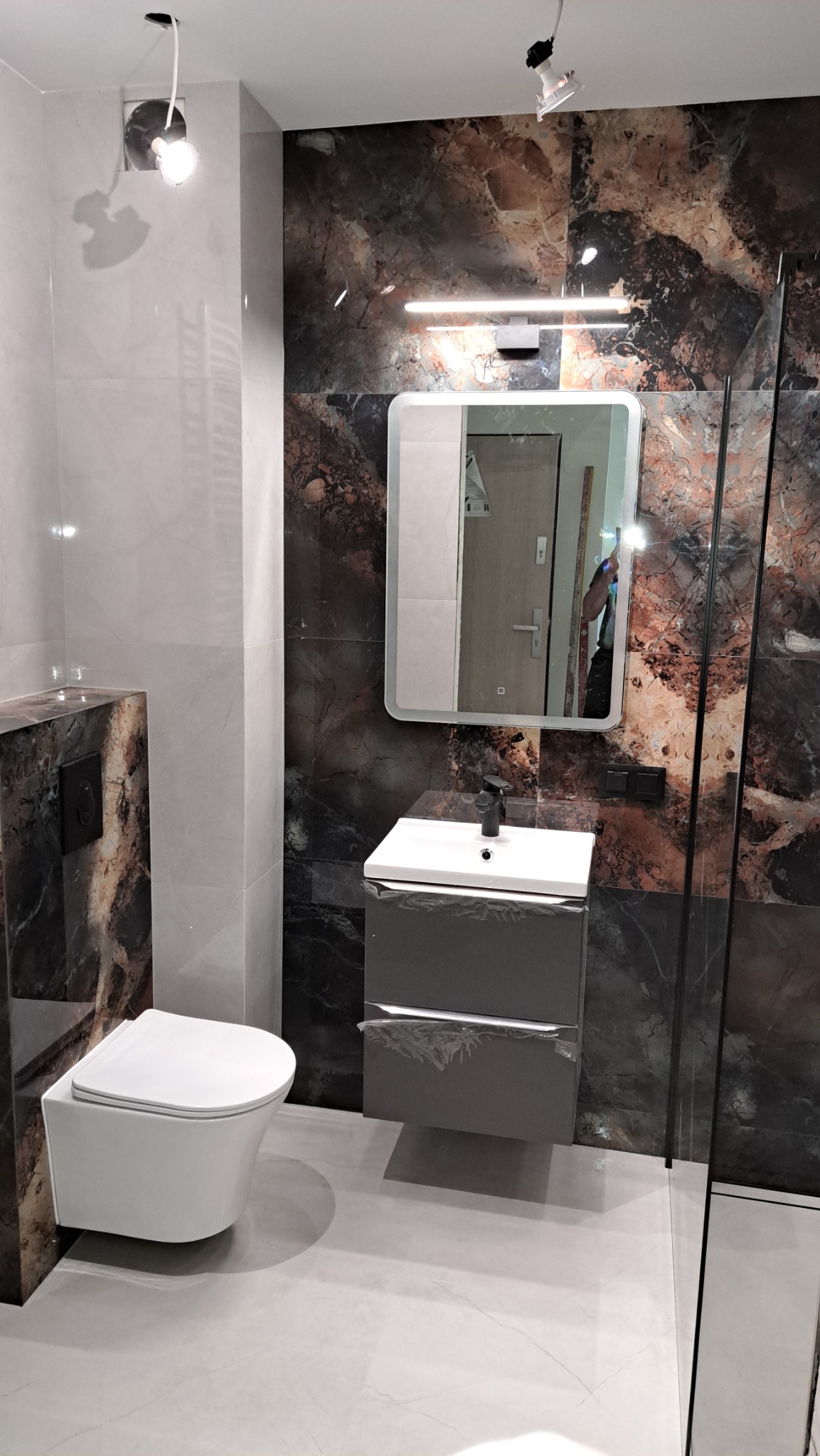 Nowoczesna łazienka z marmurowymi płytkami, podwieszaną toaletą i szafką z umywalką. Lustro z podświetleniem. Minimalistyczny design.