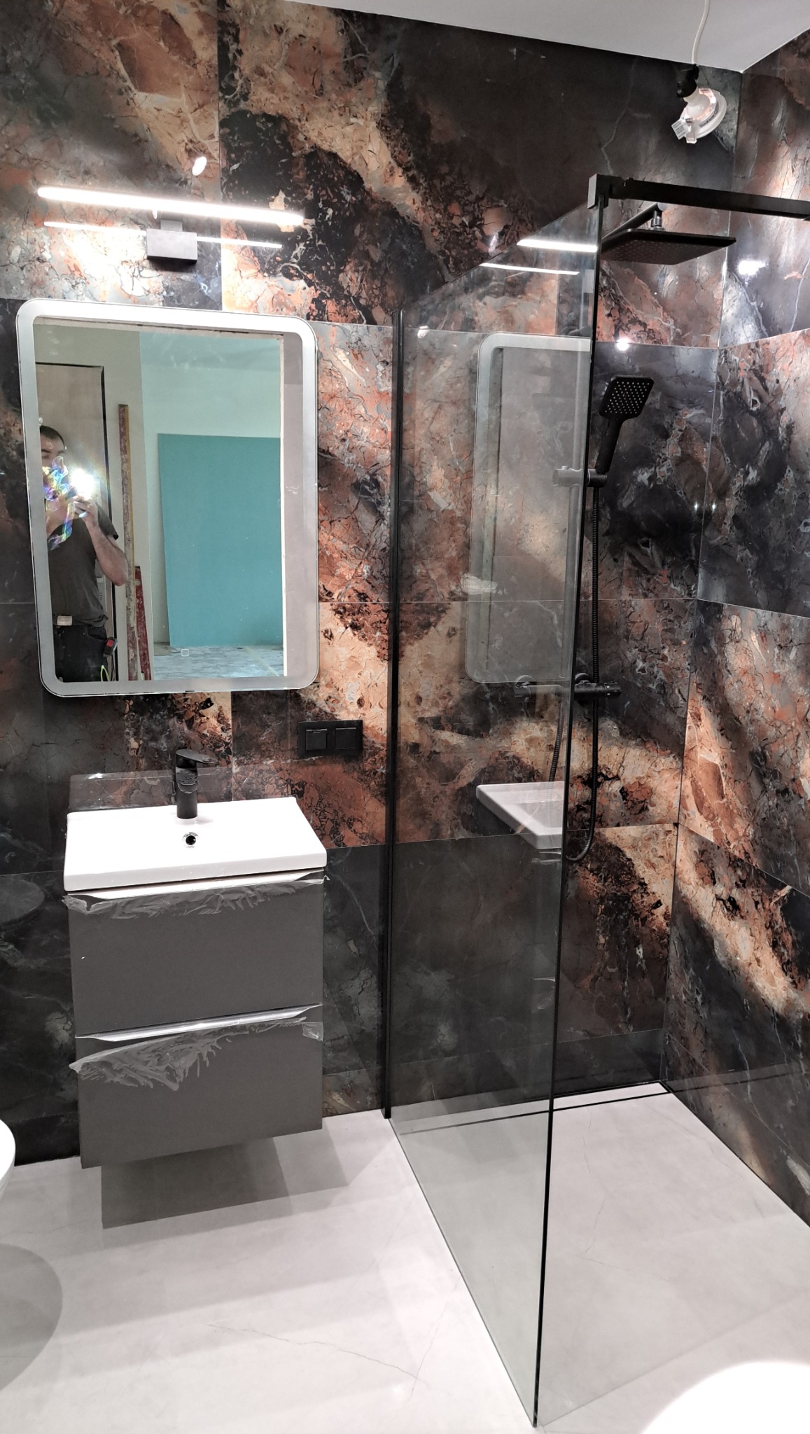 Wykończona łazienka z kabiną typu walk-in, umywalką z szafką i lustrem z podświetleniem. Ściany wyłożone płytkami imitującymi marmur w odcieniach brązu i czerni.