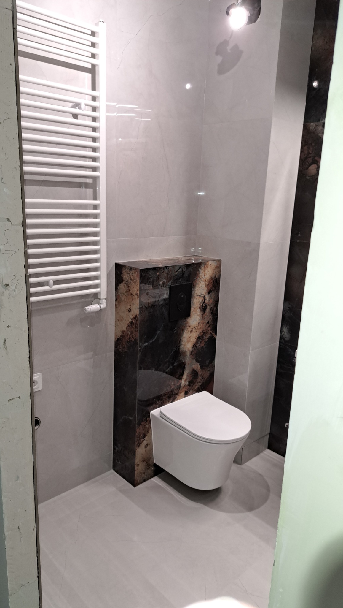 Wykończona łazienka w szarościach z podwieszaną toaletą obudowaną płytami imitującymi marmur i białym grzejnikiem drabinkowym.