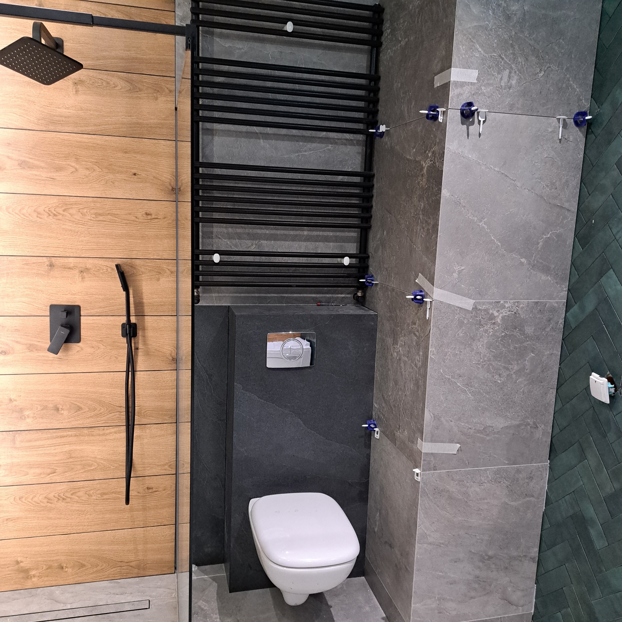 Nowoczesna łazienka w trakcie wykańczania: szare płytki imitujące kamień, drewnopodobna okładzina, czarna armatura i grzejnik. Widoczne narzędzia glazurnicze.