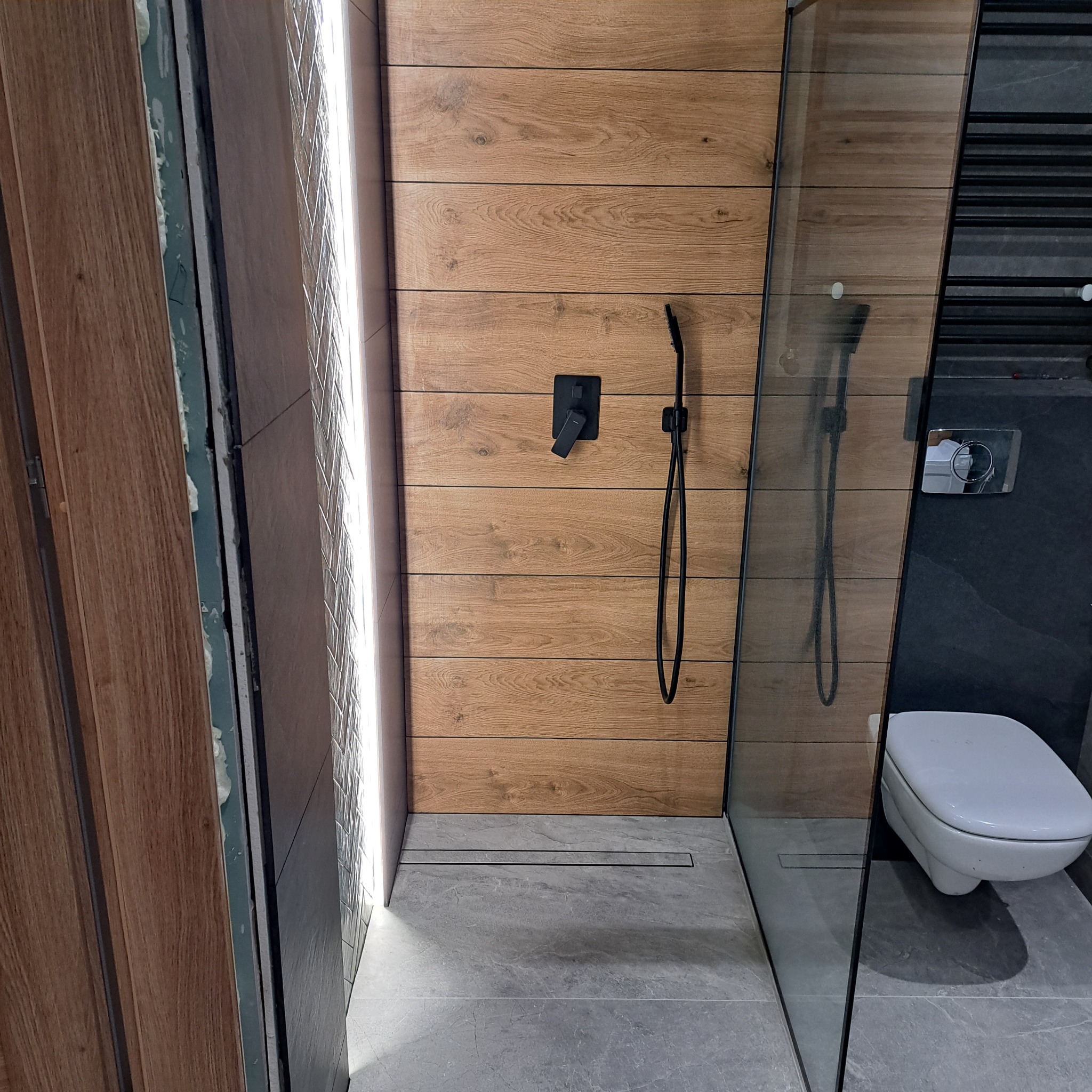 Nowoczesna kabina prysznicowa z drewnopodobnymi płytkami, czarną armaturą i odpływem liniowym. Minimalistyczny design, szklana ściana kabiny, szare płytki podłogowe.