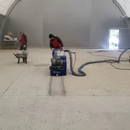 Prace przy posadzce przemysłowej w hali: szlifowanie betonu maszyną Blastrack, odkurzacz przemysłowy i pracownicy w odzieży roboczej. Widoczny fragment posadzki po szlifowaniu.