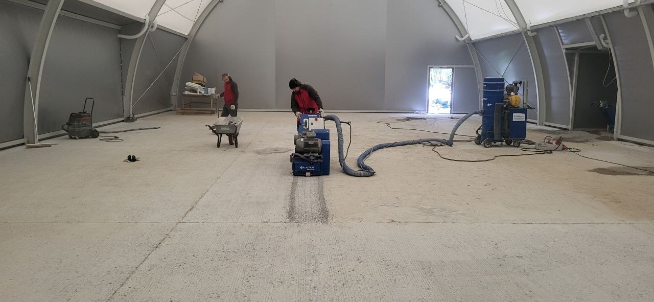 Prace przy posadzce przemysłowej w hali: szlifowanie betonu maszyną Blastrack, odkurzacz przemysłowy i pracownicy w odzieży roboczej. Widoczny fragment posadzki po szlifowaniu.