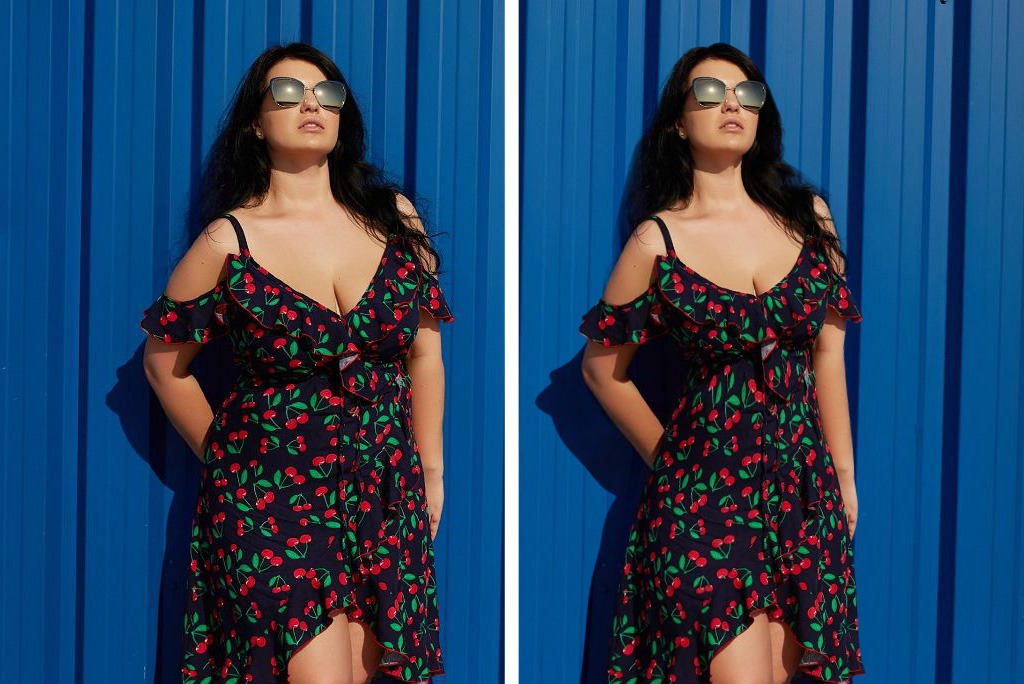 Portret kobiety w sukience z wiśniami i okularach przeciwsłonecznych, pozującej na tle niebieskiej, pionowo żłobionej ściany w słoneczny dzień.