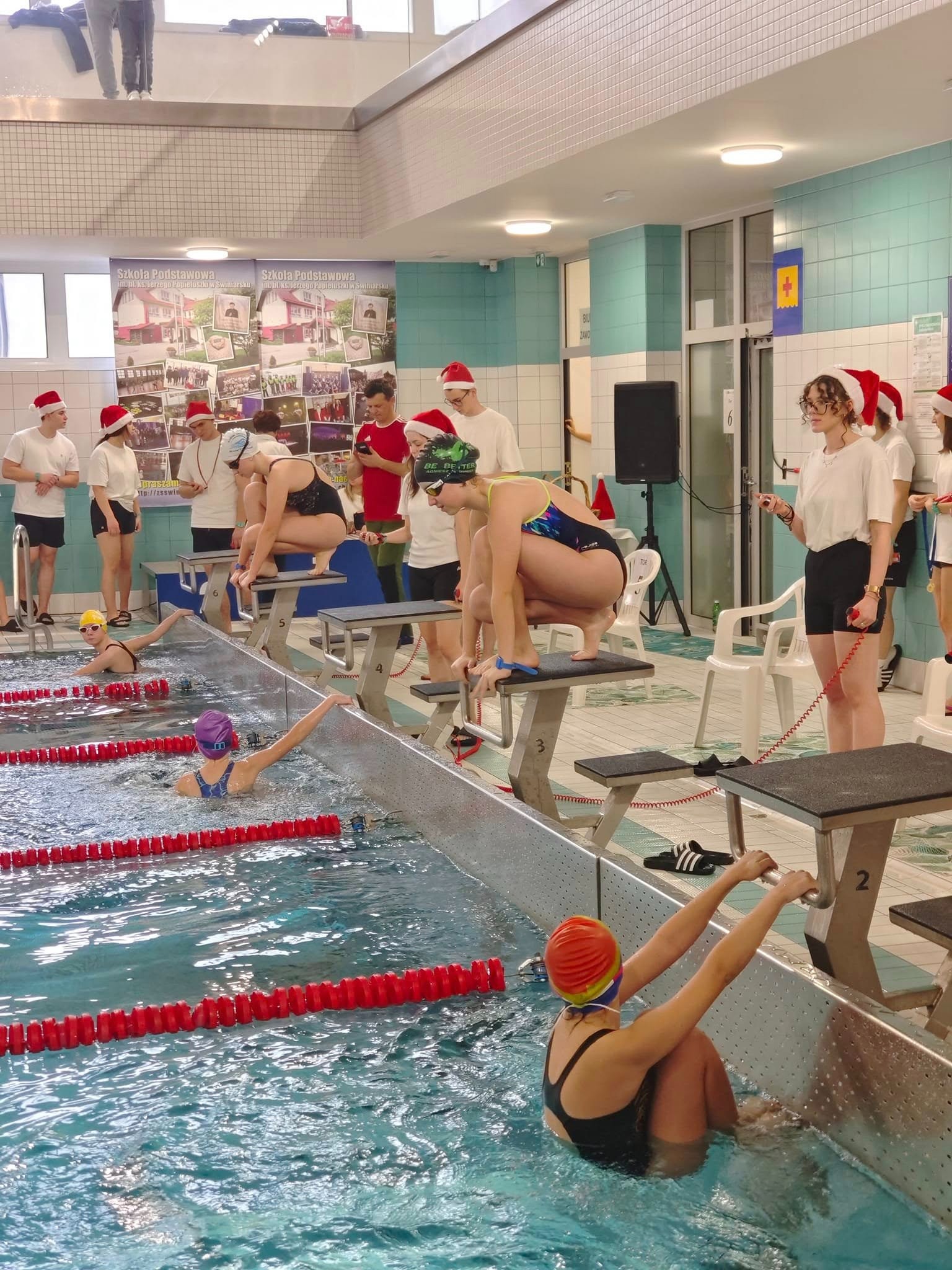 Zawody pływackie w świątecznej atmosferze: pływacy w czapkach Mikołaja na blokach startowych, oczekujący na sygnał do skoku do basenu z torami.