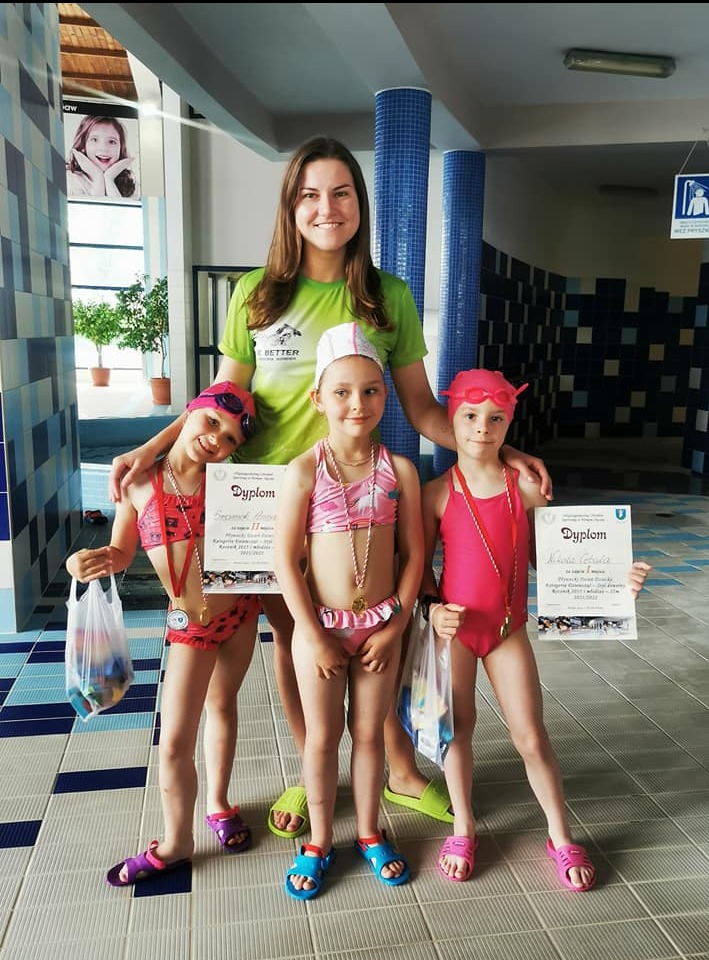 Instruktorka z uśmiechem pozuje z trzema dziewczynkami w strojach kąpielowych, które trzymają dyplomy i medale po zawodach pływackich na tle basenu.