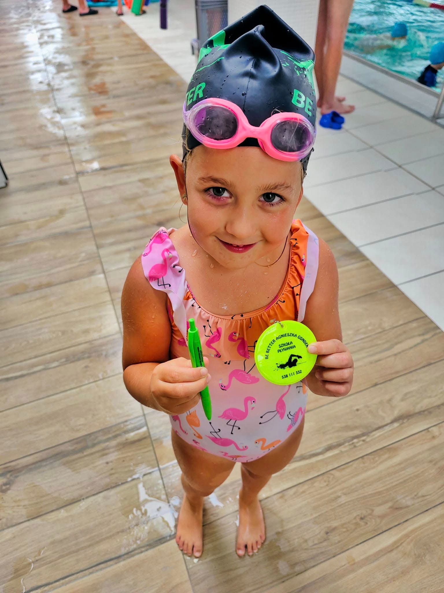 Uśmiechnięta dziewczynka w czepku i różowych okularach trzymająca zielony długopis i okrągłą odznakę szkoły pływania na tle basenu.