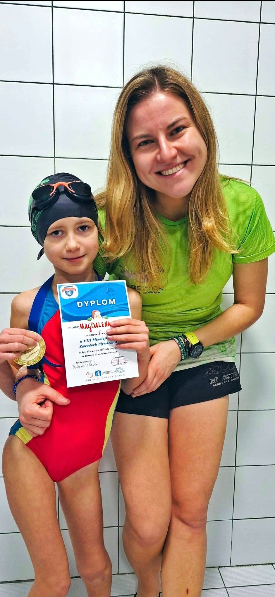 Uśmiechnięta dziewczynka w czepku i stroju kąpielowym z dyplomem i medalem, obok trenerka w sportowym stroju na tle białych płytek.