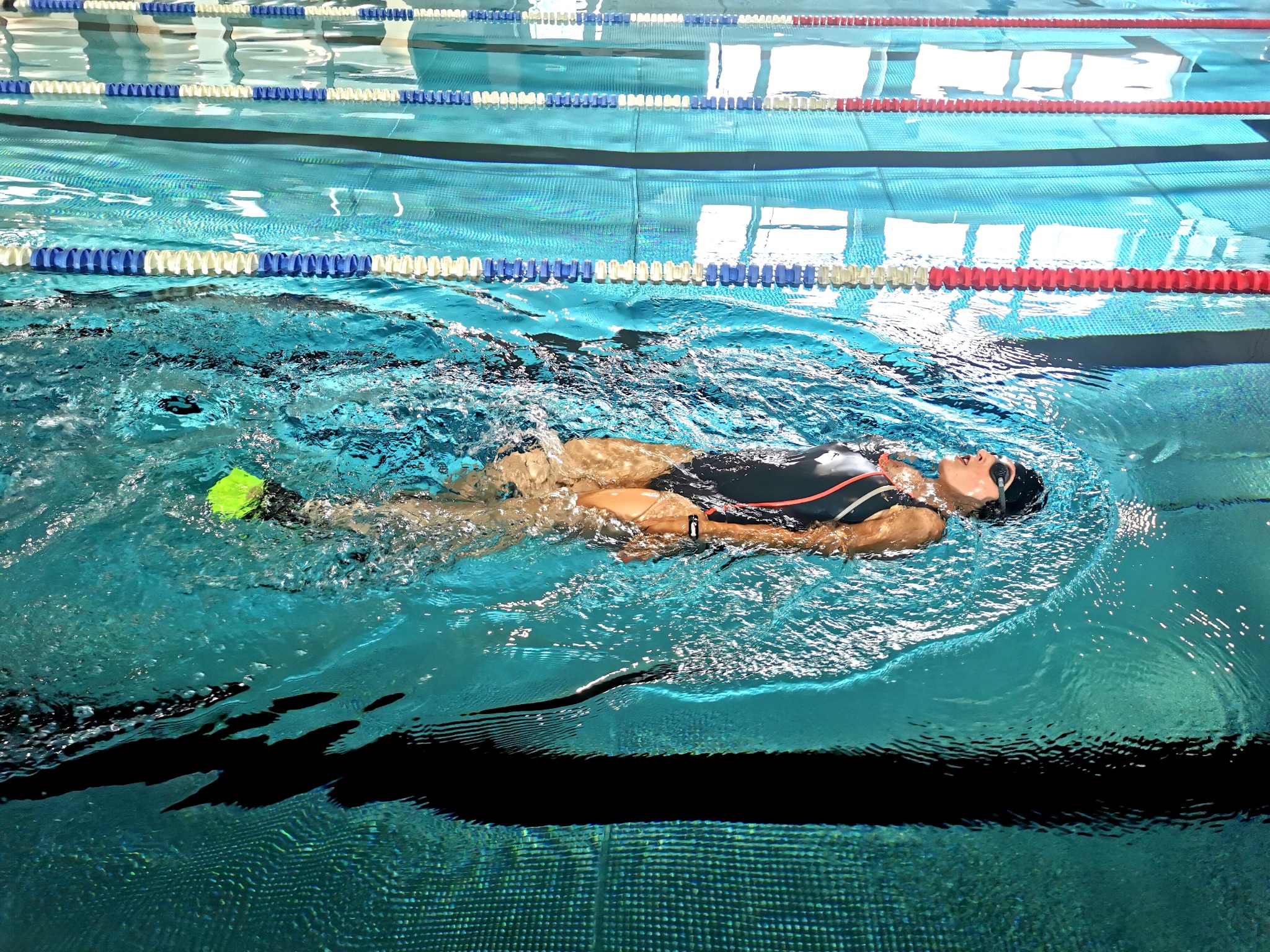 Kobieta w czepku i okularach płynie na plecach w basenie z zielonymi płetwami, wzdłuż toru oddzielonego linami z bojkami.