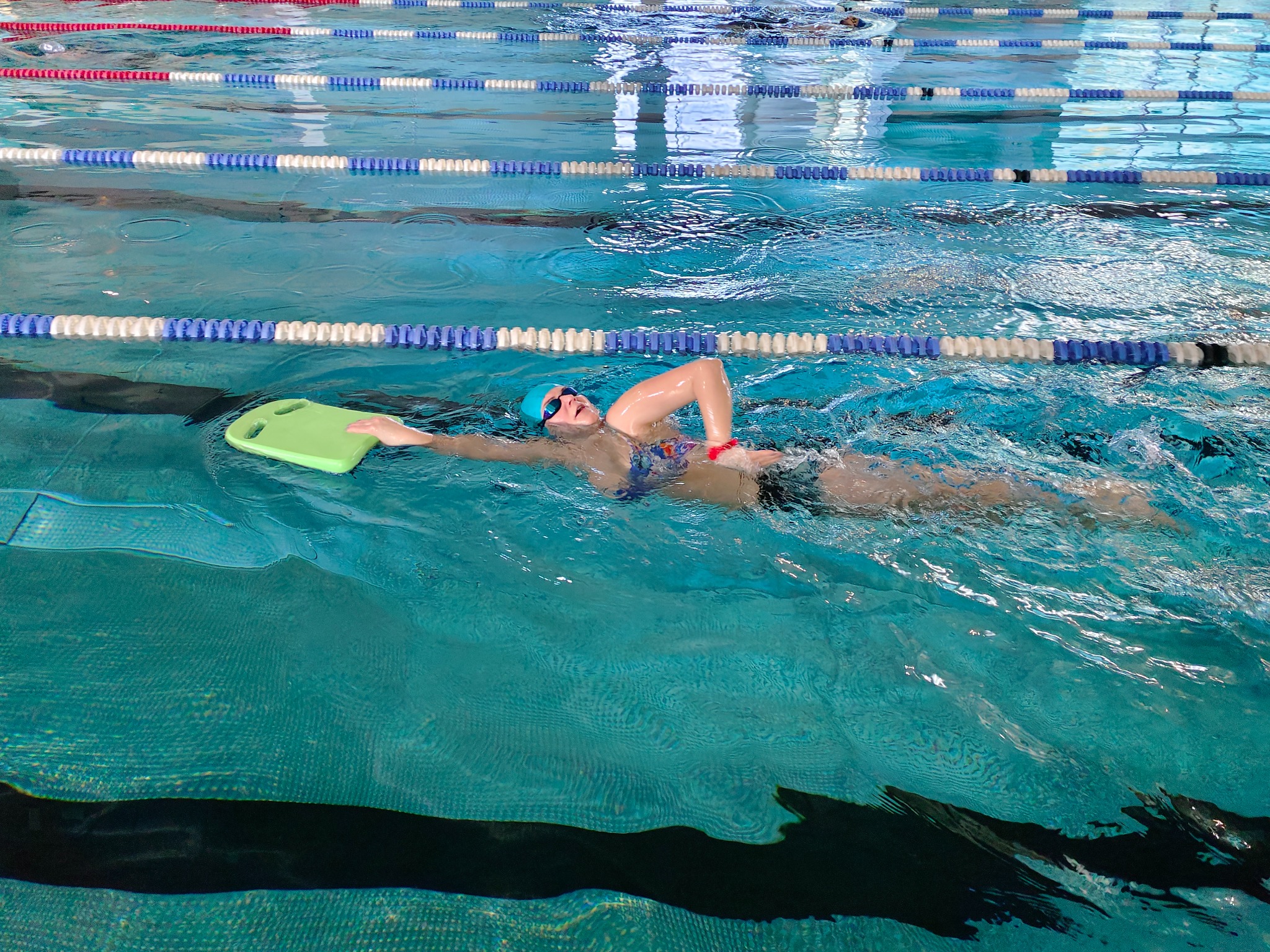 Kobieta w czepku i okularach płynie kraulem w basenie z zieloną deską, oddzielona torami z bojek. Widok z góry, woda turkusowa.