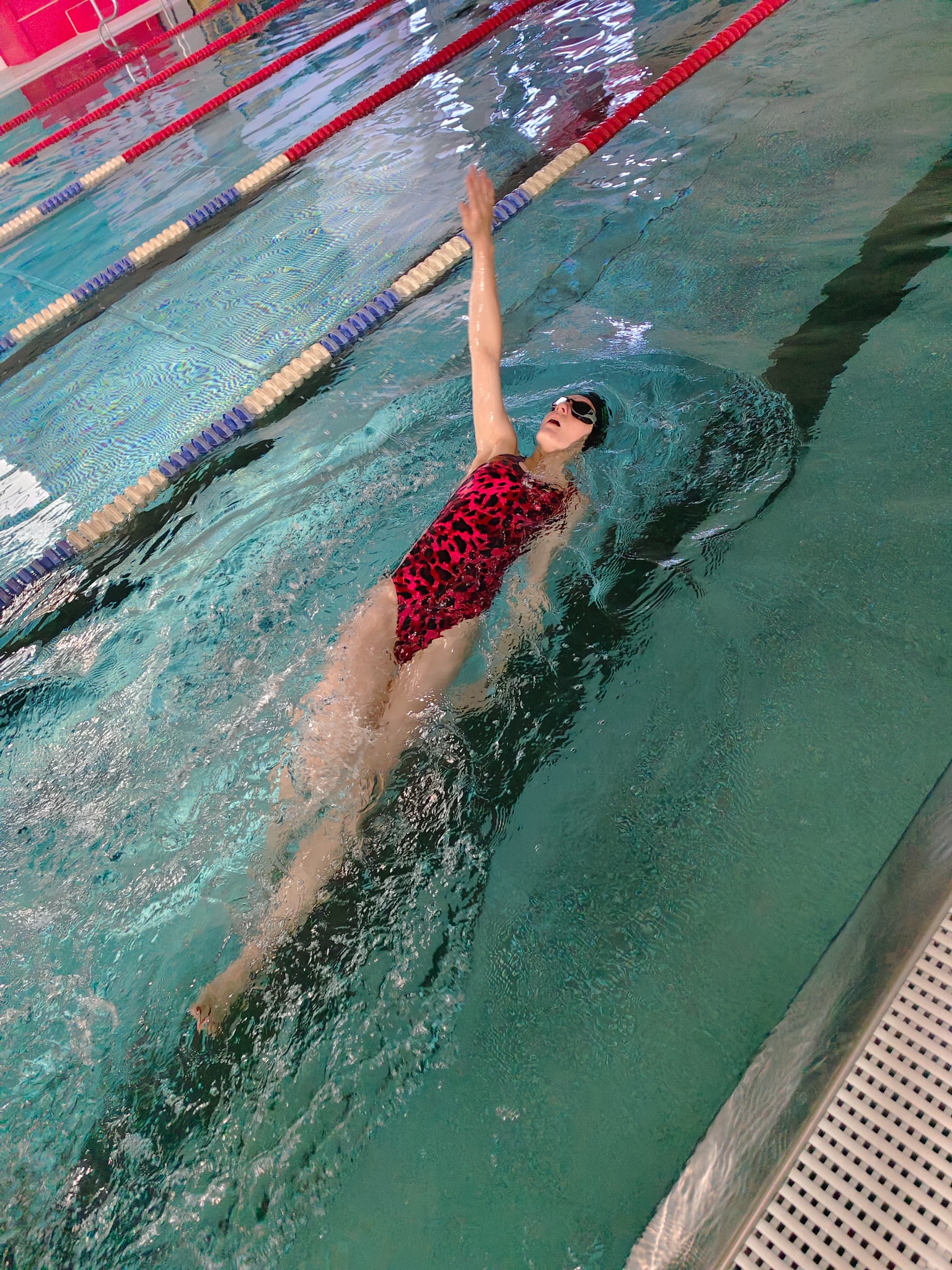 Kobieta w czepku i okularach pływa stylem grzbietowym w basenie z wyznaczonymi torami, w czerwono-czarnym kostiumie.